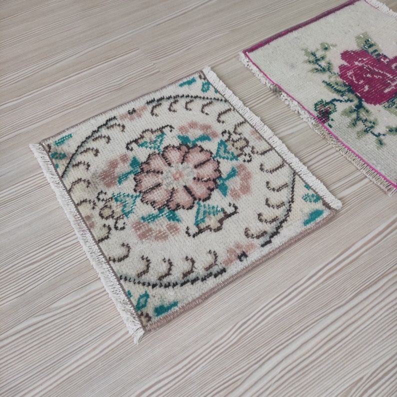 Puede incluir: Dos peque&ntilde;as alfombras cuadradas vintage con dise&ntilde;os florales. Una alfombra presenta una flor rosa central con acentos turquesa y marr&oacute;n sobre un fondo crema. La otra alfombra tiene un dise&ntilde;o floral rosa sobre un fondo crema. Ambas alfombras tienen bordes con flecos.