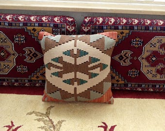 May include: A colorful patterned throw pillow with a geometric design in shades of brown, beige, and teal. The pillow is sitting on top of a red and cream patterned rug.