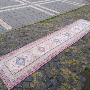 Peut inclure: Un long tapis rectangulaire avec un champ central cr&egrave;me et une bordure rose. Le tapis pr&eacute;sente des motifs g&eacute;om&eacute;triques en turquoise, rose et beige. Le tapis est pos&eacute; sur une surface pav&eacute;e.