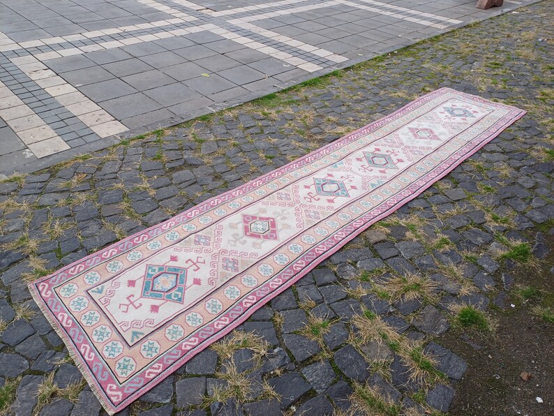 Peut inclure: Un long tapis rectangulaire avec un champ central cr&egrave;me et une bordure rose. Le tapis pr&eacute;sente des motifs g&eacute;om&eacute;triques en turquoise, rose et beige. Le tapis est pos&eacute; sur une surface pav&eacute;e.