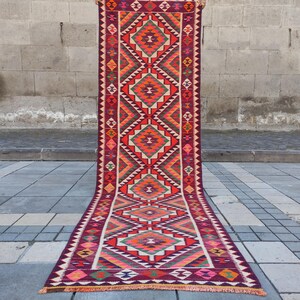 May include: A long, rectangular rug with a geometric pattern. The rug features a central design of repeating diamond shapes in shades of orange, red, green, and white, set against a deep burgundy background. The edges are lined with a zig-zag pattern.