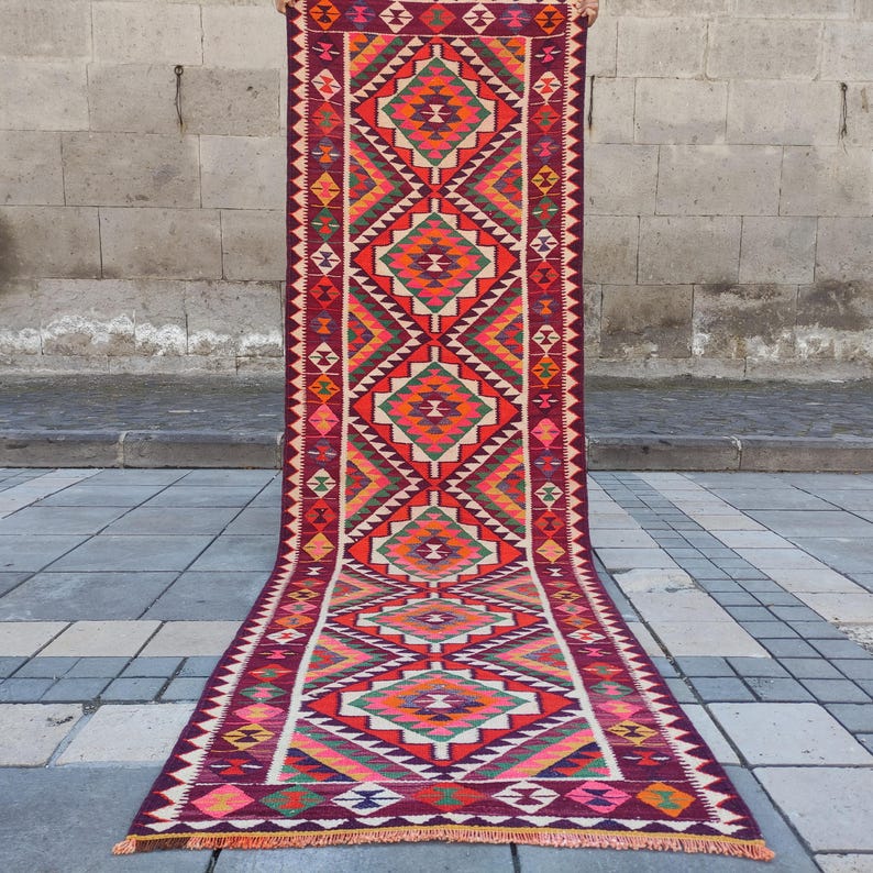 May include: A long, rectangular rug with a geometric pattern. The rug features a central design of repeating diamond shapes in shades of orange, red, green, and white, set against a deep burgundy background. The edges are lined with a zig-zag pattern.