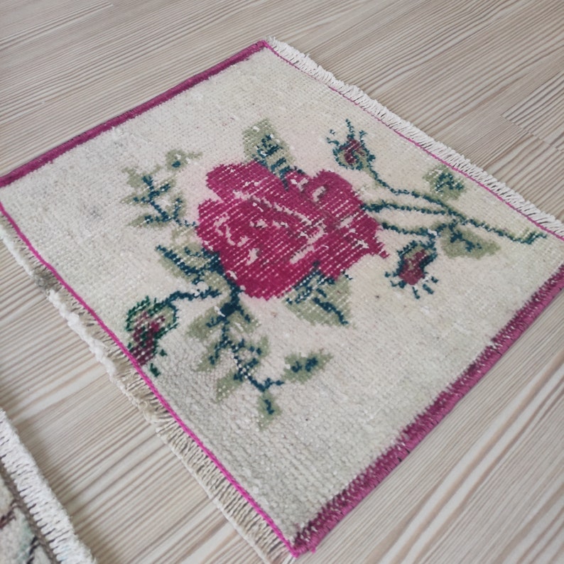 Puede incluir: Alfombra cuadrada de color crema con una gran rosa magenta en el centro. La rosa est&aacute; rodeada de hojas y capullos verdes. La alfombra tiene un borde magenta y flecos.