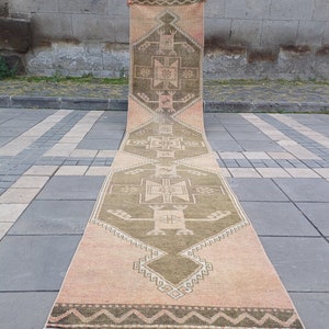 May include: A long, narrow rug with a geometric pattern in shades of beige, brown, and green. The rug has a fringe on both ends.