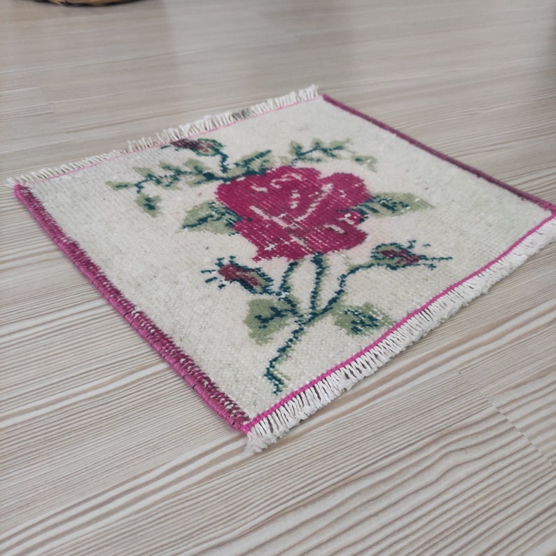 Puede incluir: Una alfombra cuadrada de color blanquecino con una gran rosa rosa vibrante en el centro. Hojas y capullos verdes rodean la rosa. La alfombra tiene un borde rosa y flecos en los bordes. La alfombra probablemente est&aacute; hecha de tela tejida.