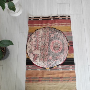 Può includere: Un cuscino rotondo con un motivo a righe con un design crema, rosso e nero si trova su un tappeto a righe colorate. Il tappeto ha una varietà di colori, tra cui rosso, arancione, giallo e marrone.