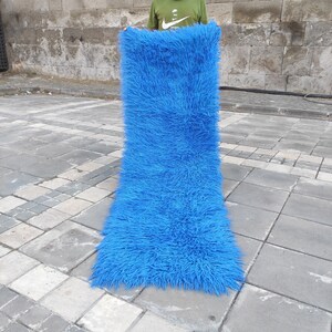 Könnte beinhalten: Ein langer, leuchtend blauer Shaggy-Teppich mit einer flauschigen Textur. Der Teppich wird von einer Person gehalten, aber die Hände der Person sind auf dem Bild nicht zu sehen.