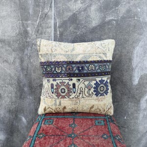 Op de afbeelding: Een vierkant, beige kussen met een patroon. Het kussen heeft een centrale band met donkerblauwe en blauwgroene bloemmotieven, met extra bloemmotieven in het onderste gedeelte. Het kussen is geplaatst op een rood en blauwgroen patroon.