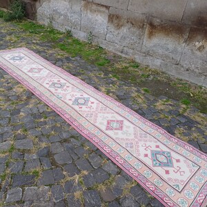 Peut inclure: Un long tapis de couloir rectangulaire avec un champ central cr&egrave;me et une bordure rose. Le tapis pr&eacute;sente des motifs g&eacute;om&eacute;triques roses, turquoise et bleu clair. Le tapis est pos&eacute; sur une surface pav&eacute;e.