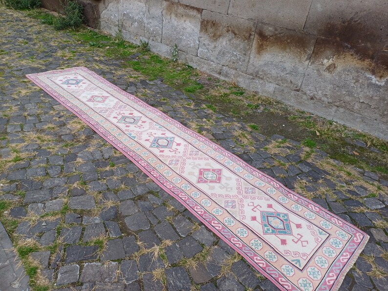 Peut inclure: Un long tapis de couloir rectangulaire avec un champ central cr&egrave;me et une bordure rose. Le tapis pr&eacute;sente des motifs g&eacute;om&eacute;triques roses, turquoise et bleu clair. Le tapis est pos&eacute; sur une surface pav&eacute;e.