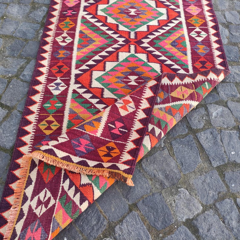 May include: A colorful, geometric patterned rug with a folded section. The rug features a burgundy background with diamond and triangle patterns in shades of orange, pink, green, and white. The edges have a fringe.