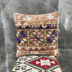 Puede incluir: Un cojín decorativo con un diseño tejido y texturizado. El cojín presenta una banda central con motivos florales y geométricos en tonos morados, rosas y verdes, sobre un fondo marrón y beige. El cojín está colocado encima de otro cojín con un patrón geométrico.