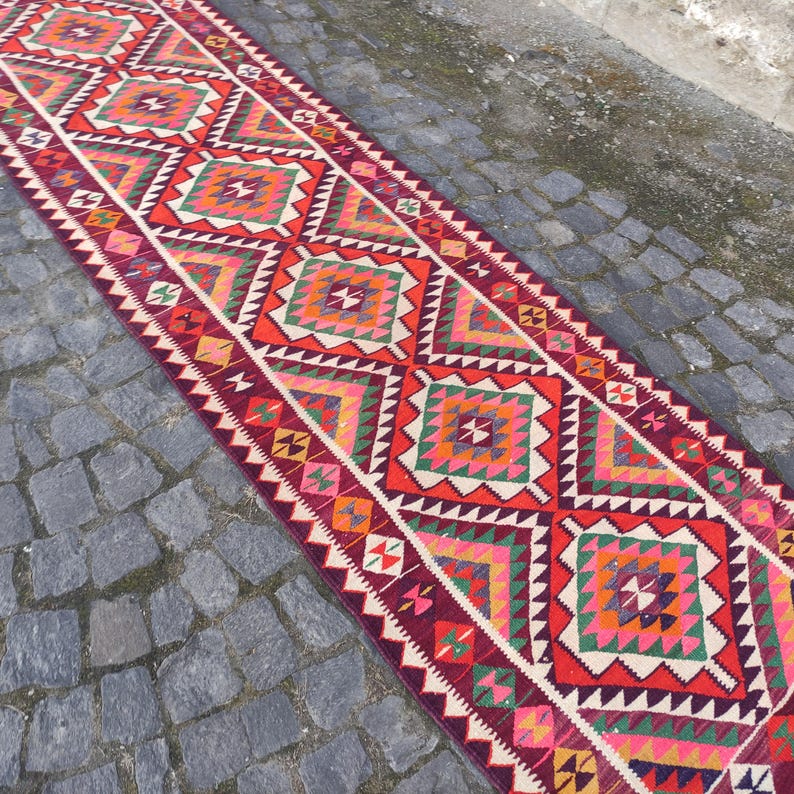 May include: A long, rectangular runner rug with a geometric pattern. The rug features a repeating design of red, orange, green, and pink squares and triangles. The edges are bordered with a white zigzag pattern.