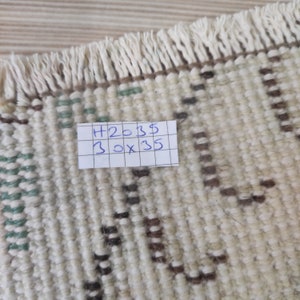 Puede incluir: Primer plano de una alfombra beige con un patr&oacute;n geom&eacute;trico en marr&oacute;n y verde. La alfombra tiene flecos en un lado. Una nota manuscrita en la alfombra dice "H2035 30 x 35". La alfombra mide aproximadamente 76 cm x 89 cm.