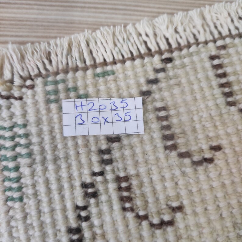 Puede incluir: Primer plano de una alfombra beige con un patr&oacute;n geom&eacute;trico en marr&oacute;n y verde. La alfombra tiene flecos en un lado. Una nota manuscrita en la alfombra dice "H2035 30 x 35". La alfombra mide aproximadamente 76 cm x 89 cm.