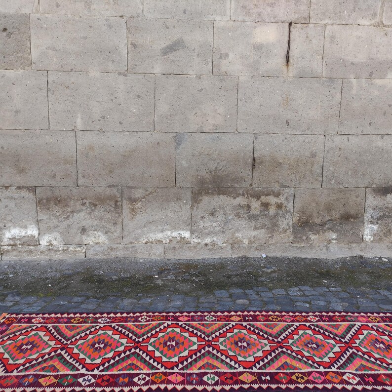 May include: A vibrant, geometric patterned rug with a red, orange, and green color scheme. The rug features diamond and zigzag designs. The rug is partially visible, with a stone wall in the background.