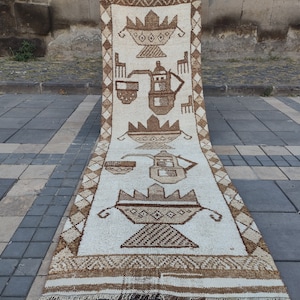 Puede incluir: Una alfombra blanca y marrón con un diseño geométrico que presenta un motivo repetido de una tetera y tazas. La alfombra está doblada por la mitad y se encuentra sobre una superficie de baldosas grises.
