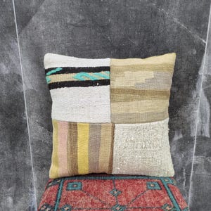 May include: Square patchwork pillow with a variety of textures and colors. The pillow features four distinct sections: white with black and turquoise stripes, beige, striped with pink and yellow, and textured off-white.
