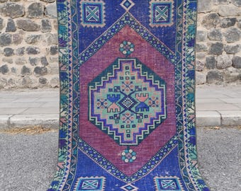 Könnte beinhalten: Ein rechteckiger Webteppich mit einem großen zentralen Rautenmotiv in tiefem Magenta, umgeben von einem dunkelblauen Feld. Das Design umfasst komplexe geometrische Muster in Petrol, Rosa und Weiß mit einer dekorativen Bordüre aus sich wiederholenden Formen.