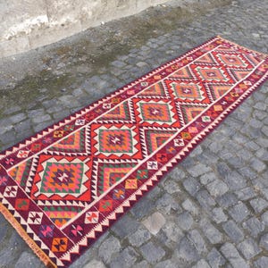 May include: A long, rectangular, hand-woven rug with a geometric pattern. The rug features a repeating diamond design in shades of red, orange, green, and white. The edges are finished with a fringe. The rug is displayed on a cobblestone surface.