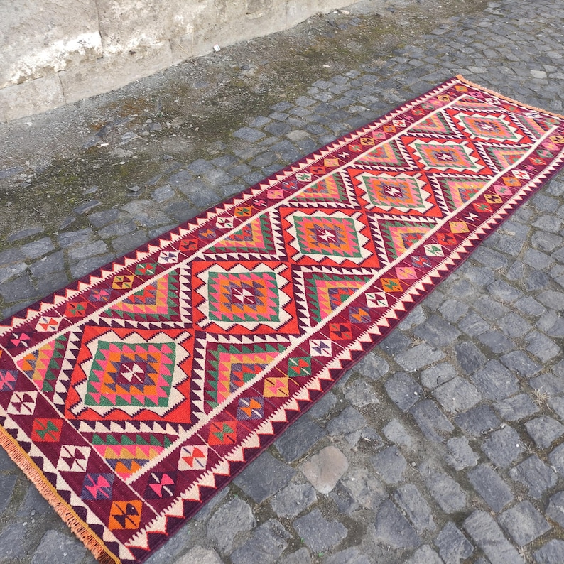 May include: A long, rectangular, hand-woven rug with a geometric pattern. The rug features a repeating diamond design in shades of red, orange, green, and white. The edges are finished with a fringe. The rug is displayed on a cobblestone surface.