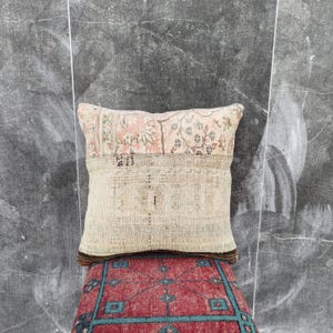 May include: A rectangular decorative pillow with a beige, pink, and brown patterned design. The pillow is placed on a red rug with a geometric pattern. The pillow has a vintage, distressed look.