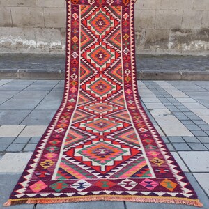 May include: A long, rectangular runner rug with a geometric pattern. The rug features a central design of diamonds and triangles in shades of red, orange, green, and pink, set against a deep burgundy background. The edges are finished with a fringe.