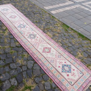 Peut inclure: Un long tapis rectangulaire avec un champ central cr&egrave;me et un motif g&eacute;om&eacute;trique r&eacute;p&eacute;titif dans les tons de rose, bleu et vert. Le tapis a une bordure rose et est pos&eacute; sur une surface pav&eacute;e.
