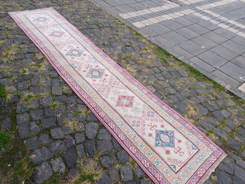 Peut inclure: Un long tapis rectangulaire avec un champ central cr&egrave;me et un motif g&eacute;om&eacute;trique r&eacute;p&eacute;titif dans les tons de rose, bleu et vert. Le tapis a une bordure rose et est pos&eacute; sur une surface pav&eacute;e.