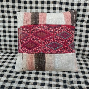 Può includere: Un cuscino decorativo rettangolare con un motivo geometrico. La sezione centrale presenta un motivo a rombi rossi, affiancato da sezioni a righe nei toni del rosa, marrone e crema. Il cuscino è in tessuto.