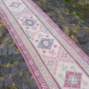 Peut inclure: Un tapis de couloir rectangulaire avec un motif g&eacute;om&eacute;trique central dans les tons de rose, turquoise et beige. Le tapis a une bordure avec un motif r&eacute;p&eacute;titif et est pos&eacute; sur une surface pav&eacute;e. Le design du tapis est sym&eacute;trique.
