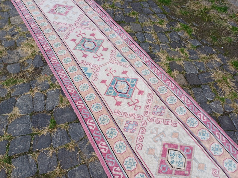 Peut inclure: Un tapis de couloir rectangulaire avec un motif g&eacute;om&eacute;trique central dans les tons de rose, turquoise et beige. Le tapis a une bordure avec un motif r&eacute;p&eacute;titif et est pos&eacute; sur une surface pav&eacute;e. Le design du tapis est sym&eacute;trique.