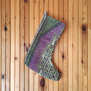 Può includere: Un calzino di colore viola, verde e bianco con un motivo tessuto. Il calzino è appeso su uno sfondo di pannello di legno.