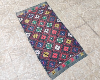 May include: A colorful rug with a geometric pattern in shades of red, green, blue, and yellow. The rug has a fringed edge and is made of woven fabric.