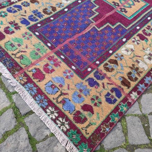 May include: A vintage Turkish rug with a beige background and a colorful floral pattern. The rug has a burgundy border with green, blue, and white accents. The rug has a fringe on all sides.