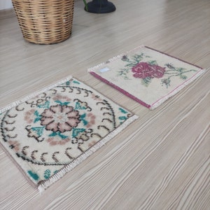 Puede incluir: Dos peque&ntilde;as alfombras cuadradas tejidas a mano con dise&ntilde;os florales. Una presenta una rosa rosa, la otra un motivo floral central en rosa y turquesa. Ambas tienen bordes con flecos y est&aacute;n sobre un suelo de madera clara.