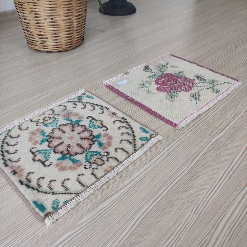 Puede incluir: Dos peque&ntilde;as alfombras cuadradas tejidas a mano con dise&ntilde;os florales. Una presenta una rosa rosa, la otra un motivo floral central en rosa y turquesa. Ambas tienen bordes con flecos y est&aacute;n sobre un suelo de madera clara.