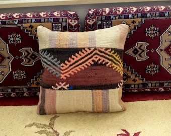 May include: A colorful, handwoven throw pillow with a geometric pattern in shades of red, orange, brown, and beige. The pillow is sitting on a cream-colored rug with a floral design.