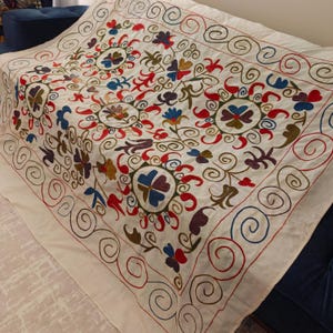 May include: Cream-coloured textile with detailed embroidery. The design includes floral and swirling patterns in red, blue, green, and brown. The edges are bordered with a repeating spiral design. This textile is likely a decorative throw or wall hanging.