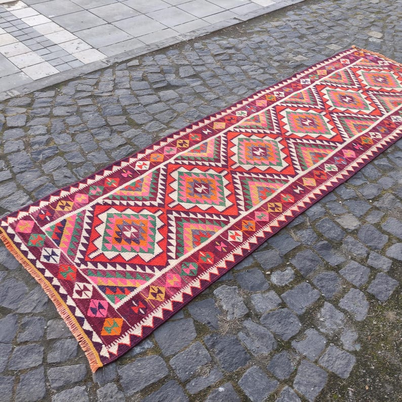 May include: A long, rectangular runner rug with a geometric pattern. The rug features a repeating design of diamonds and zigzags in shades of red, orange, pink, green, and white, set against a dark burgundy background. The rug has a fringed edge.