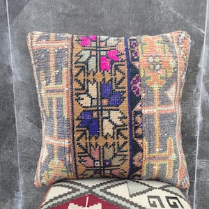 Può includere: Un cuscino quadrato decorativo con un design intrecciato e fantasia. Il cuscino presenta un pannello centrale con motivi floreali e geometrici nei toni del marrone, blu, rosa e arancione. I bordi hanno un bordo grigio e arancione. Il cuscino è posto sopra un altro cuscino fantasia.