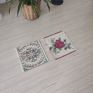 Puede incluir: Dos peque&ntilde;as alfombras cuadradas vintage con dise&ntilde;os florales. Una presenta una flor rosa central con hojas verdes, la otra tiene un patr&oacute;n circular con acentos rosas y turquesas. Ambas tienen bordes con flecos y est&aacute;n sobre una superficie de madera clara.
