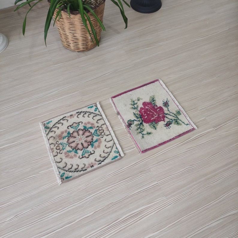 Puede incluir: Dos peque&ntilde;as alfombras cuadradas vintage con dise&ntilde;os florales. Una presenta una flor rosa central con hojas verdes, la otra tiene un patr&oacute;n circular con acentos rosas y turquesas. Ambas tienen bordes con flecos y est&aacute;n sobre una superficie de madera clara.