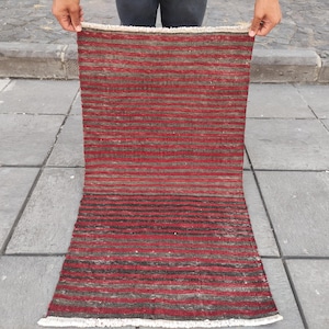 May include: A rectangular woven rug featuring horizontal stripes in shades of deep red, dark brown, and black. The textile has a textured appearance with a visible weave pattern and short, off-white fringe detailing along the top and bottom edges.