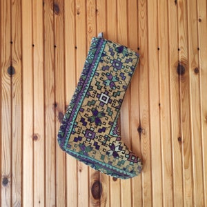 Può includere: Una calza di Natale decorativa con un motivo geometrico nei toni del beige, viola, verde e verde acqua. La calza è appesa a una parete di legno. Il motivo include quadrati, diamanti e forme floreali.