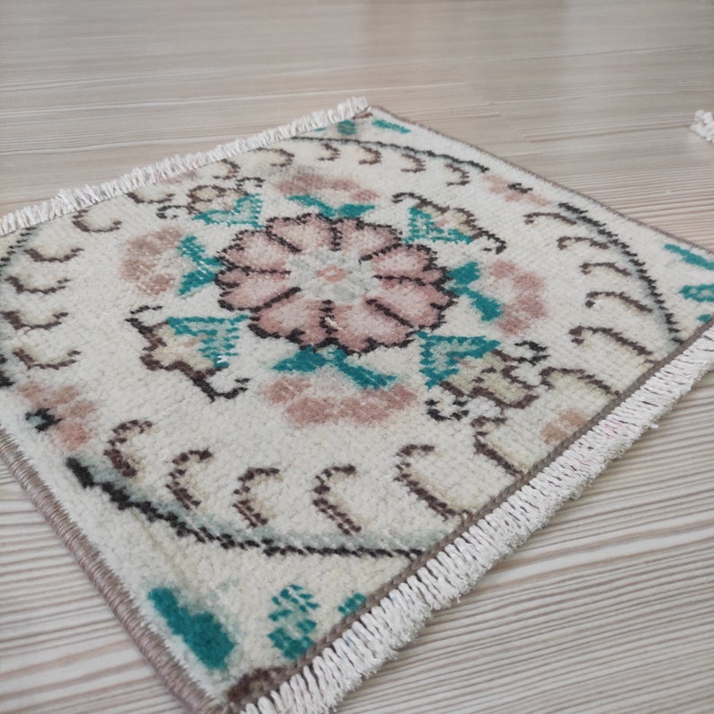 Puede incluir: Alfombra cuadrada de color blanco roto con dise&ntilde;o floral. La alfombra presenta una flor rosa central rodeada de detalles turquesa y marr&oacute;n. Los bordes est&aacute;n adornados con flecos. La alfombra es probablemente hecha a mano.