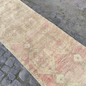 Puede incluir: Una alfombra alargada de tipo pasillero con una paleta de colores rosa y crema desvaídos. El tejido presenta un patrón geométrico repetitivo de grandes medallones centrales con bordes angulares intrincados y pequeños motivos de rombos.