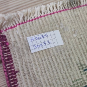 Puede incluir: Primer plano de una peque&ntilde;a alfombra blanquecina con un tejido texturizado. La alfombra tiene un borde rosa y flecos en un borde. Una etiqueta con el texto manuscrito "H2035 30x37" (76cm x 94cm) est&aacute; adjunta.