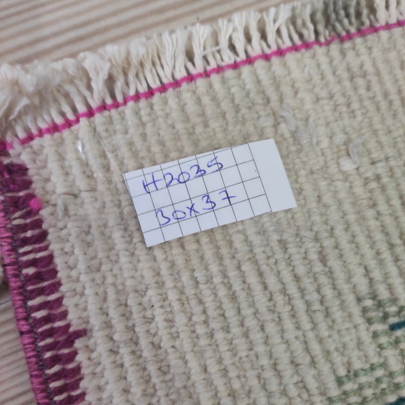 Puede incluir: Primer plano de una peque&ntilde;a alfombra blanquecina con un tejido texturizado. La alfombra tiene un borde rosa y flecos en un borde. Una etiqueta con el texto manuscrito "H2035 30x37" (76cm x 94cm) est&aacute; adjunta.