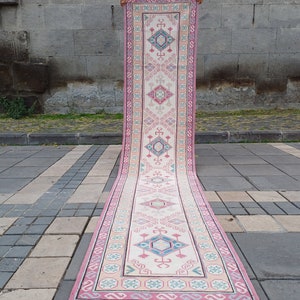 Peut inclure: Un long tapis rectangulaire avec un champ central cr&egrave;me et un motif g&eacute;om&eacute;trique r&eacute;p&eacute;titif en rose, bleu et beige. Le tapis a une bordure rose et est expos&eacute; &agrave; l'ext&eacute;rieur sur une surface en pierre.